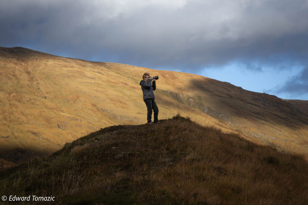 A photographer stands on a grassy hill, capturing the surrounding golden mountains under moody clouds.
