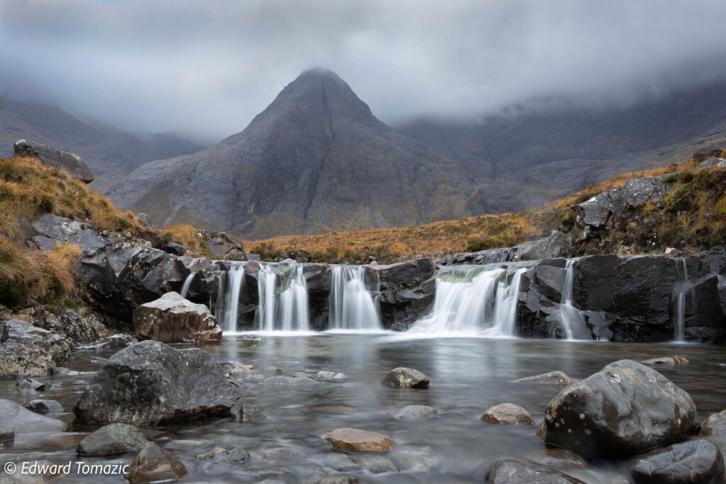 A misty mountain waterfall flows into a rocky pool beneath a dramatic peak under low cloud.