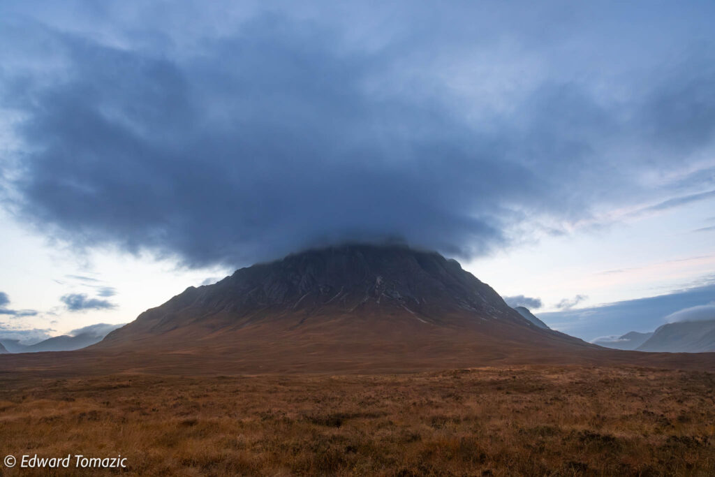 A solitary mountain rises from open moorland, its summit concealed by a dense, dark cloud.