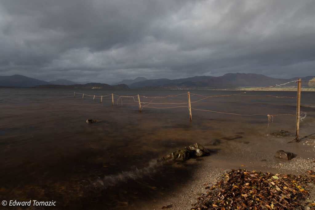 A moody lakeside scene with a partially submerged fence, dark storm clouds, gentle waves, and distant hills under dramatic light.