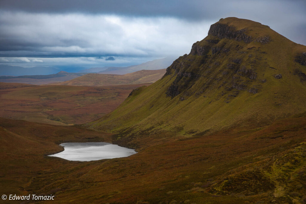 A small loch sits beneath a steep green hillside in the Quiraing, with rolling moorland and heavy clouds stretching into the distance.