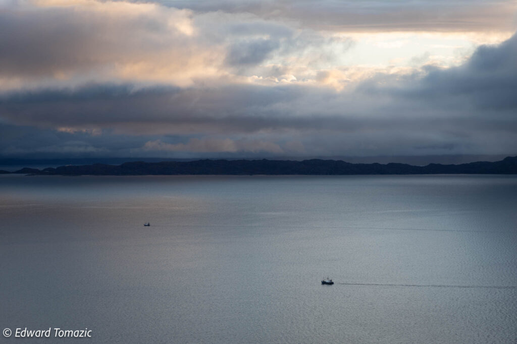 Two small boats move across calm water beneath a dramatic, layered sky along the Scottish coast.