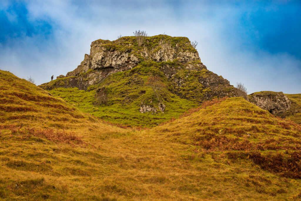 A rugged grassy hill formation rises under a cloudy sky, with a small figure standing on the left ridge in the Fairy Glen of the Isle of Skye.
