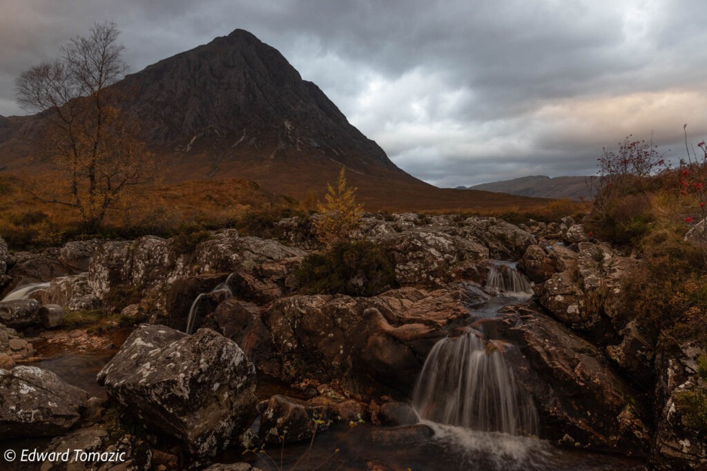 Water cascades over rocky foreground beneath a dramatic mountain and stormy sky in Glencoe, Scotland.