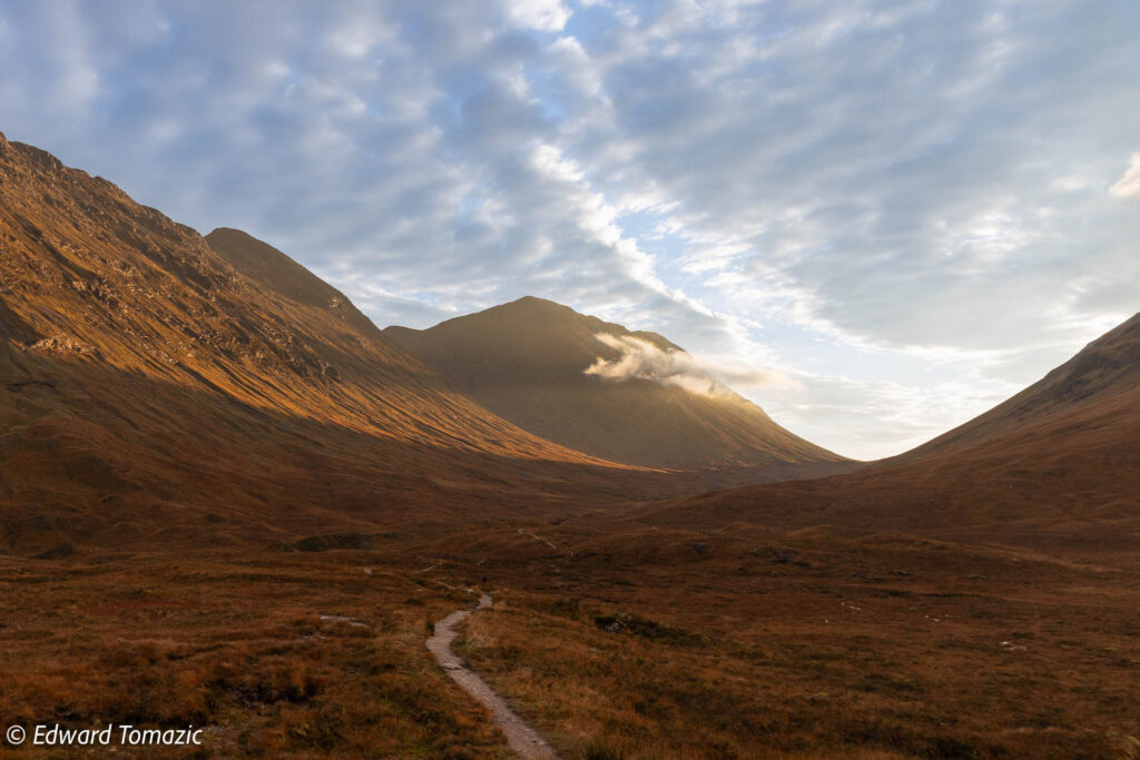 A winding path leads through a golden-brown valley toward sunlit mountains under a textured, cloud-filled sky.