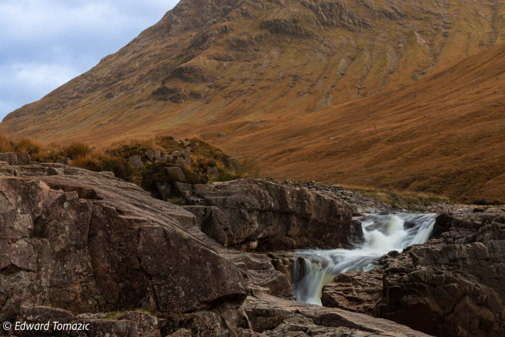 Small waterfall flowing through rocky terrain beneath a rugged mountain in the Scottish Highlands under a cloudy sky.