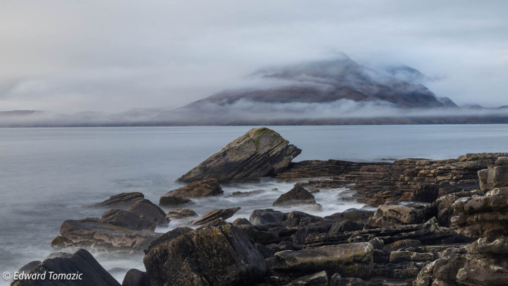 Fog drifts across a rocky coastal shoreline with calm water and mist-covered mountains in the background.