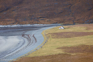 A solitary cottage beside a curving tidal shoreline beneath muted brown hills in a remote coastal landscape.