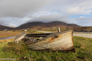 An abandoned wooden boat resting in grassy marshland beneath cloud-covered mountains on the Isle of Skye, Scotland.