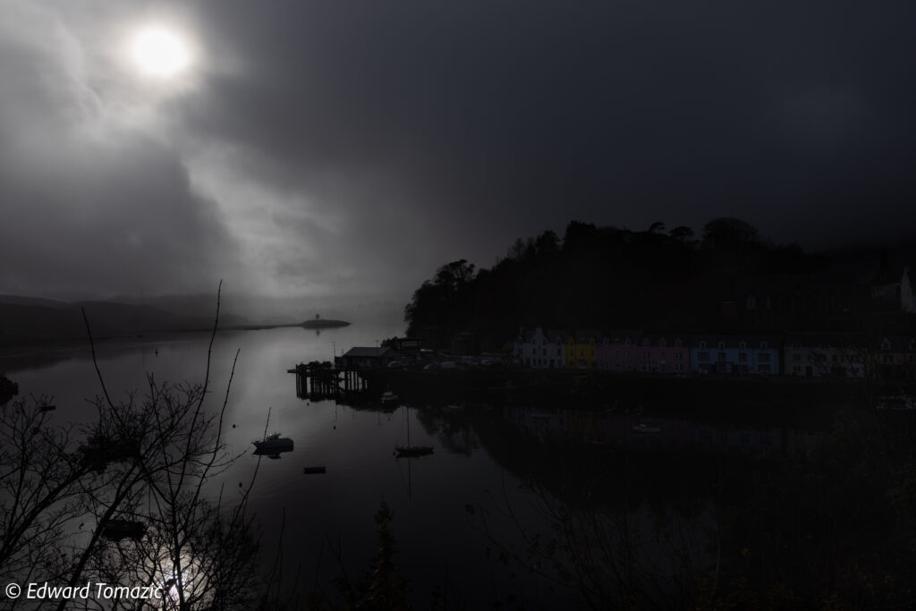 Dark and atmospheric harbor scene with storm clouds, misty light, reflective water, and colorful coastal houses partially illuminated beneath a dramatic Scottish sky.